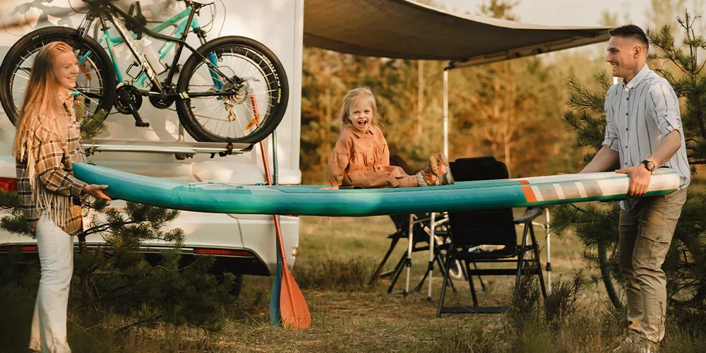 Parents lifting a paddleboard with their child sitting on it, parked beside a caravan with bikes attached.