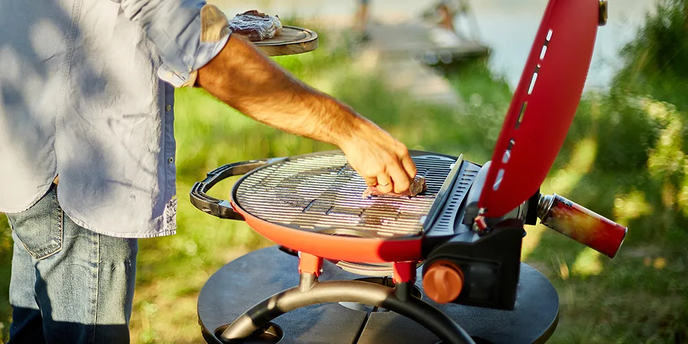 Person cooking on a portable red BBQ grill beside a peaceful lakeside setting.