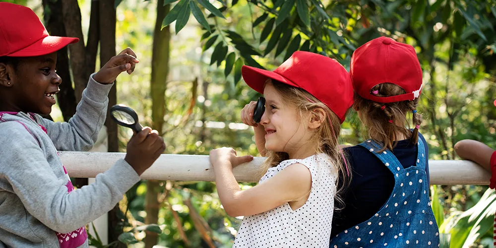 Three young children exploring outdoors with magnifying glasses and red caps, smiling and observing nature.