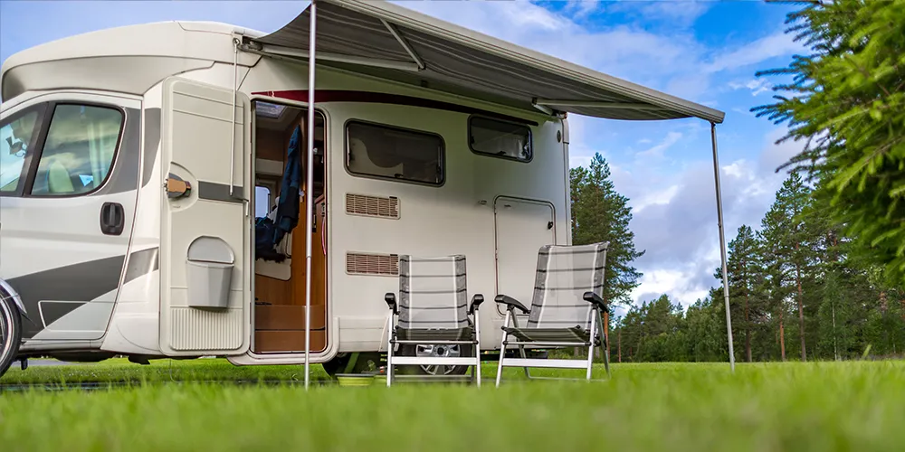 Parked motorhome with two empty camp chairs under the awning, set on a quiet grassy site surrounded by trees.