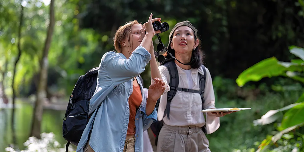 Two women wildlife watching in a lush forest, one pointing while the other holds binoculars and a field guide, both wearing backpacks.