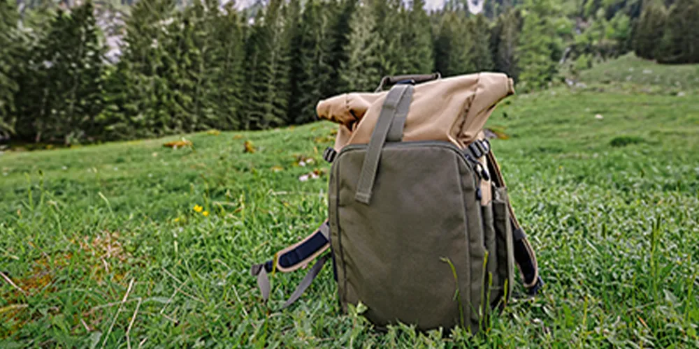 Lightweight roll-top backpack sitting on a grassy meadow with forested mountains in the distance.