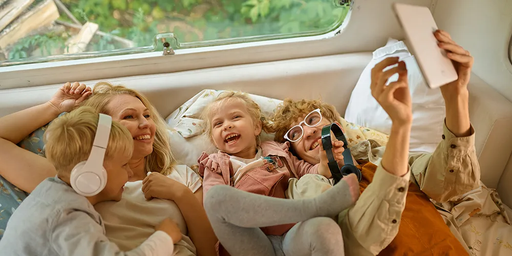 A young girl holding binoculars playfully inside a bright, cozy caravan while her family smiles and watches.