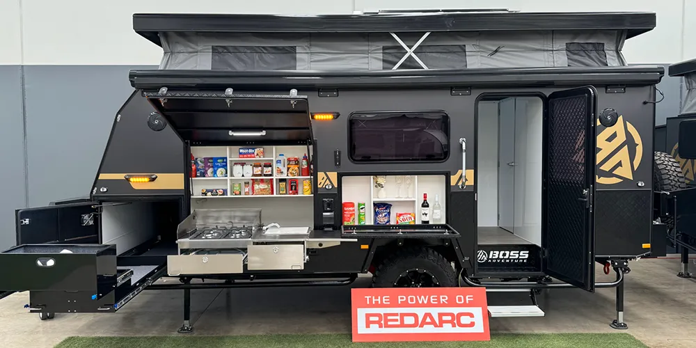 Side view of the Boss Adventure Barossa 16 off road hybrid caravan with its pop top roof raised, outdoor kitchen and pantry fully extended, and a red sign reading “The Power of Redarc” in front.