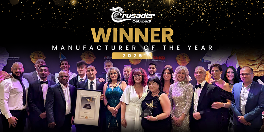 **Alt text:** Group photo of the Crusader Caravans team in formal attire, posing on stage with a framed certificate and star-shaped trophy beneath a black and gold banner that reads “Crusader Caravans – WINNER – Manufacturer of the Year 2025.”
