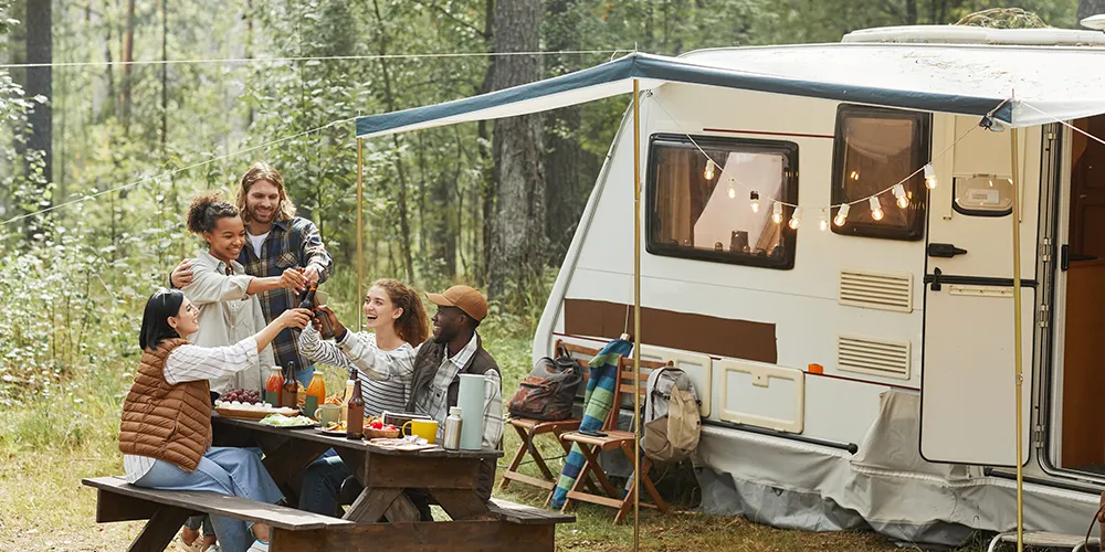 Group of friends enjoying a meal and toasting drinks at a picnic table outside a caravan in a forest campsite, with string lights adding a festive touch.