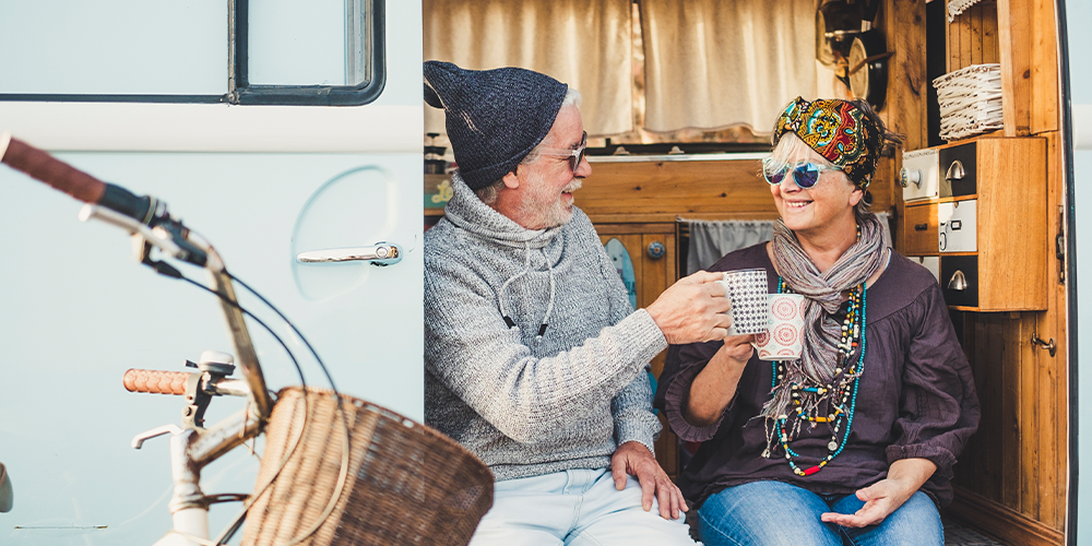 Senior couple enjoying a warm drink together outside their caravan, smiling and relaxed during a road trip.