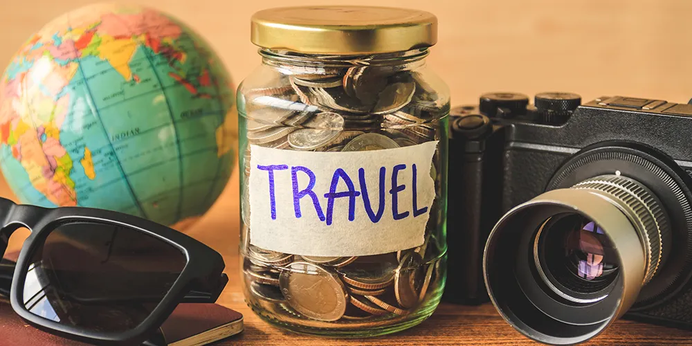 Glass jar labeled "Travel" filled with coins, surrounded by a globe, camera, sunglasses, and a passport symbolising saving money for travel adventures.